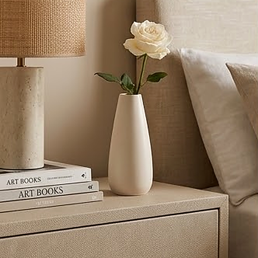 Slim off white ceramic vase with a single white rose on a bedroom nightstand beside a rattan shade lamp