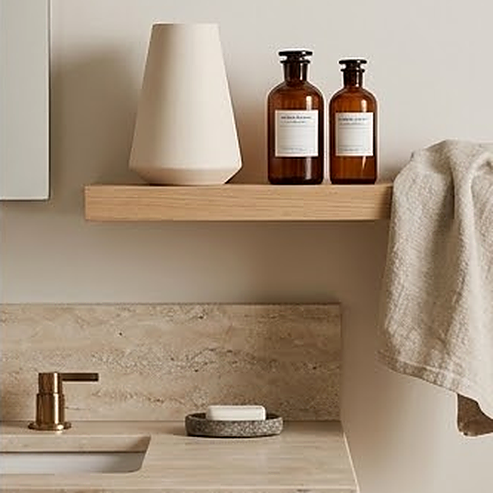 Off white ceramic vase on a floating oak shelf in a travertine bathroom beside amber glass bottles