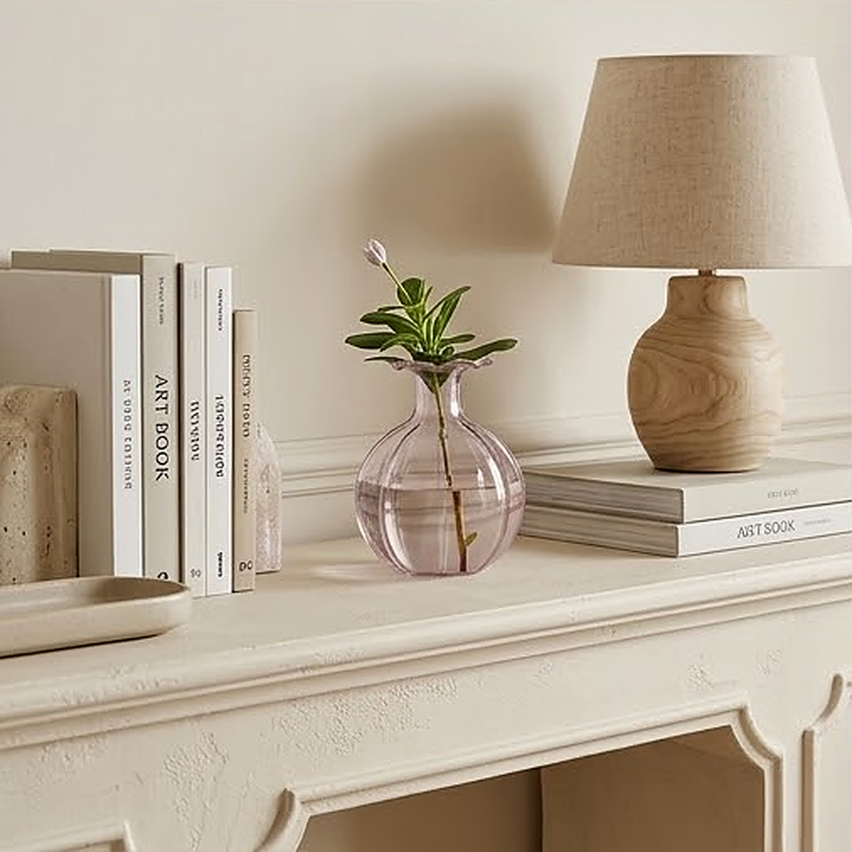 The pink glass vase holding a single green sprig on a white mantel, next to books and a wooden lamp