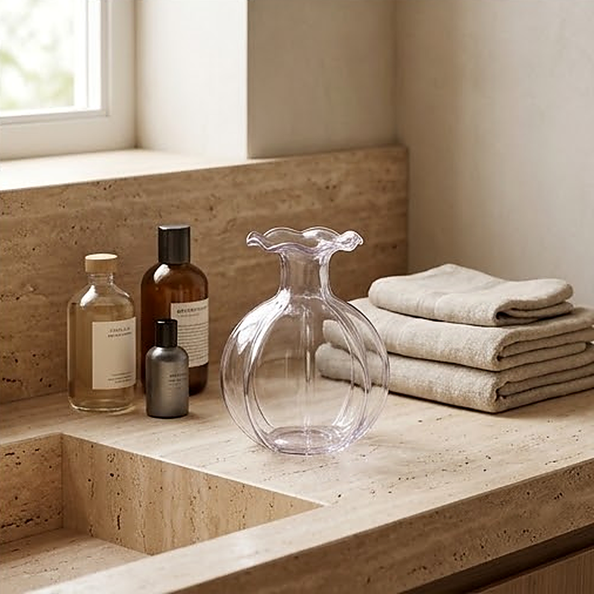 The pink glass vase on a travertine bathroom counter beside amber bottles and folded towels, reading almost clear in the light