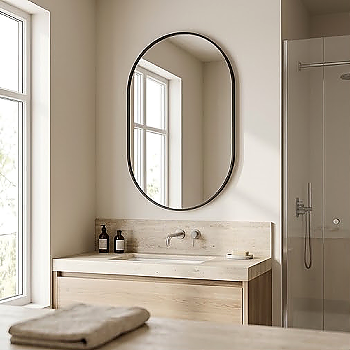 Black framed oval wall mirror hung vertically above a pale stone bathroom vanity beside a walk in shower